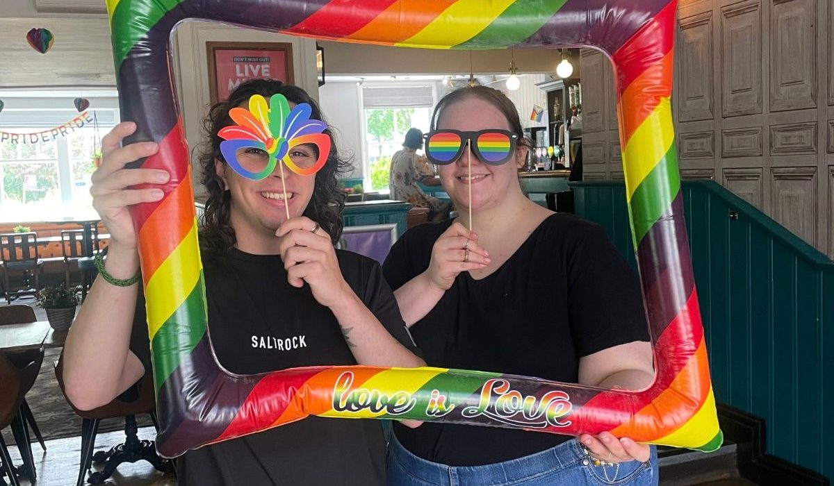Two people posing with a large inflatable rainbow pride frame reading "love is love" at an indoor Pride event. Both wear black t-shirts and pride-themed accessories including rainbow sunglasses and a rainbow fan. The venue is a pub or bar with wooden paneling and green wainscoting, decorated with Pride signage and rainbow decorations visible in the background.