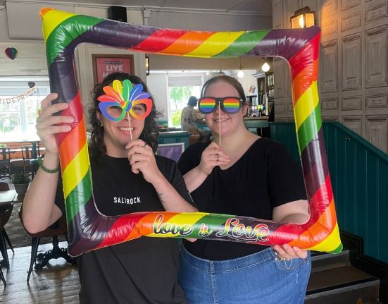 Two people posing with a large inflatable rainbow pride frame reading "love is love" at an indoor Pride event. Both wear black t-shirts and pride-themed accessories including rainbow sunglasses and a rainbow fan. The venue is a pub or bar with wooden paneling and green wainscoting, decorated with Pride signage and rainbow decorations visible in the background.
