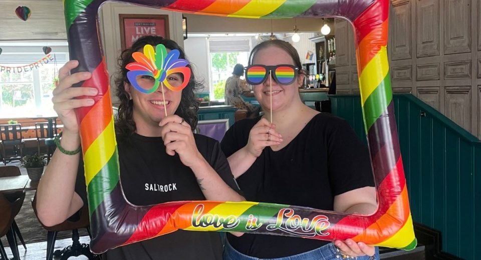 Two people posing with a large inflatable rainbow pride frame reading "love is love" at an indoor Pride event. Both wear black t-shirts and pride-themed accessories including rainbow sunglasses and a rainbow fan. The venue is a pub or bar with wooden paneling and green wainscoting, decorated with Pride signage and rainbow decorations visible in the background.