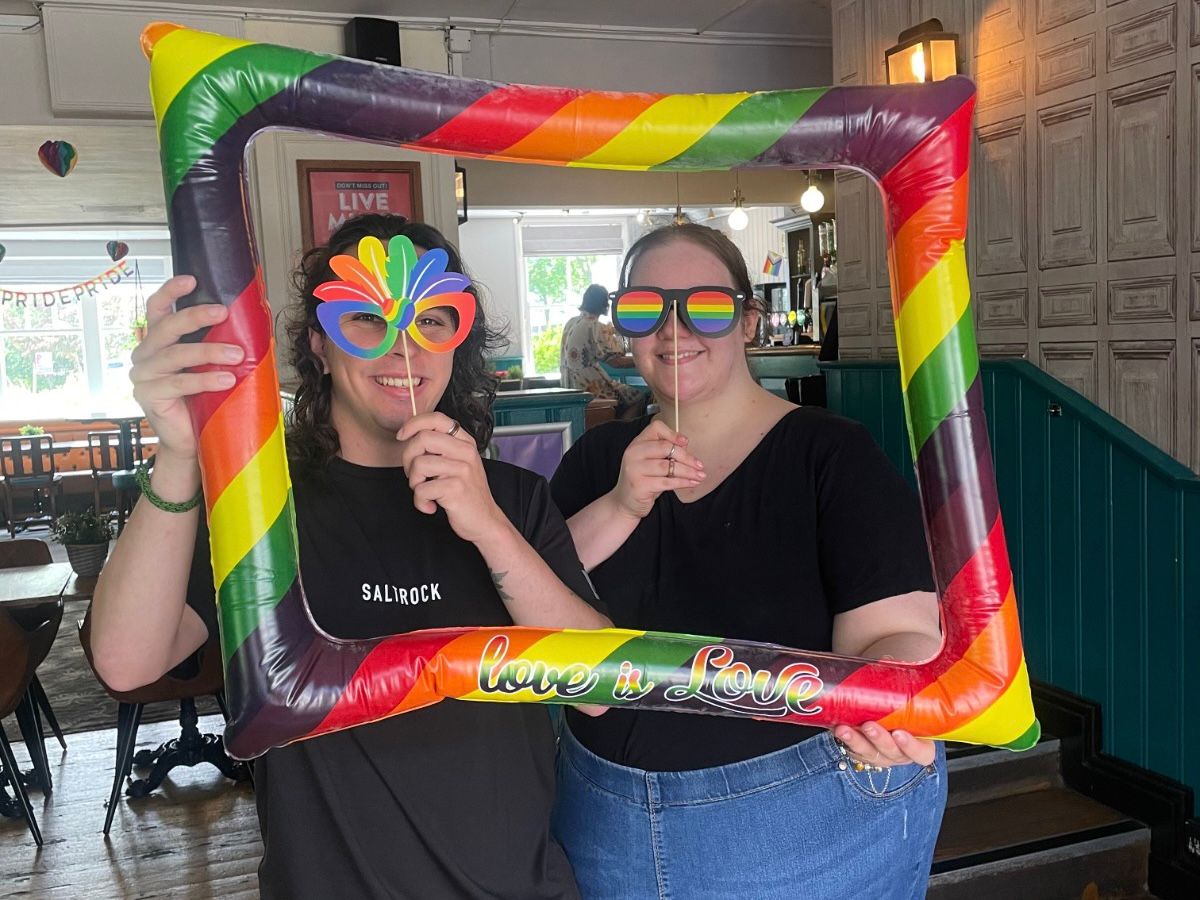 Two people posing with a large inflatable rainbow pride frame reading "love is love" at an indoor Pride event. Both wear black t-shirts and pride-themed accessories including rainbow sunglasses and a rainbow fan. The venue is a pub or bar with wooden paneling and green wainscoting, decorated with Pride signage and rainbow decorations visible in the background.