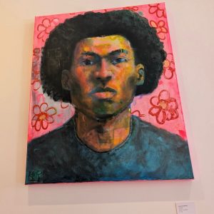 A pop art style portrait painting featuring a person rendered in vibrant colors including yellows, oranges, blues, and greens. The background is bright pink with red decorative floral patterns bordering the composition. The figure wears a dark-colored shirt. A small placard is displayed below the painting on a gallery wall.