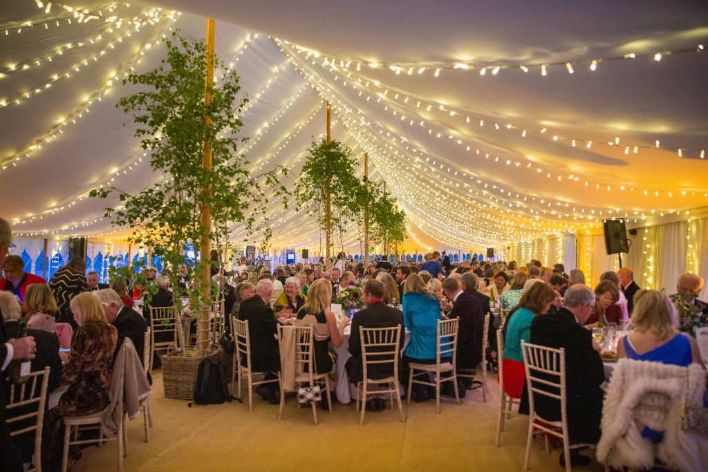Large indoor gala event at Chettle House with numerous guests in evening wear gathered in an elegantly decorated marquee. Warm string lights drape across the white fabric ceiling, illuminating the space in golden tones. Tall ivy-covered wooden poles, round tables with place settings, and white chairs are arranged throughout the venue. The atmosphere is festive and formal, with guests mingling and dining. Photography by David Lewis.