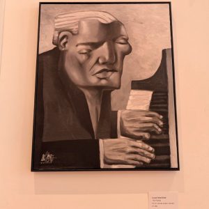 A modernist-style painting in muted beige and brown tones showing a profile view of a figure's head tilted downward, with hands visible holding geometric architectural shapes. The artwork is framed with a black border and displayed on a gallery wall. A placard below identifies the work as "The Planner" by Lucas Greenfield, oil on canvas board, framed, priced at £1,500.