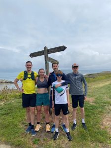 Five people in athletic wear stand in front of a coastal path signpost at Ferry Bridge, Langton Herring, Dorset, with sea and countryside visible behind them