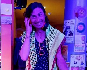 A person smiling at the camera in a neon-lit venue bathed in purple, pink, and orange lighting. They wear layered clothing including a patterned shirt and have sunglasses hanging around their neck. Behind them are posters and event notices on the wall, including "Conscious Rhythms" and "Pop-Up in Dorchester" signage