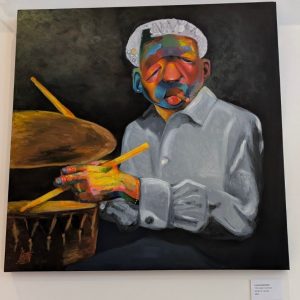 A portrait painting depicting a figure with a white headwrap and a face painted in vibrant colors including oranges, blues, reds, and greens. The figure wears a light gray shirt and holds colorful paintbrushes in hands that are also painted in bright hues. Paint containers sit at the lower left of the composition against a dark background. A placard below identifies the work by Lucas Greenfield, titled "The Colour Drummer," oil on canvas.
