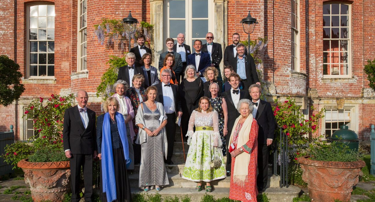 Formal group portrait of approximately 30 people in evening wear, including Annabel Elliot (Queen Camilla's sister) and other prominent Dorset residents, arranged in three rows in front of a red brick Georgian building with a white-columned central entrance. The group includes members of Dorset County Hospital Charity’s Emergency and Critical Care Appeal committee. Climbing wisteria and flowering plants frame the doorway, with terracotta urns positioned on either side of the steps. Photography by David Lewis.