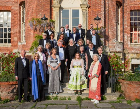 Formal group portrait of approximately 30 people in evening wear, including Annabel Elliot (Queen Camilla's sister) and other prominent Dorset residents, arranged in three rows in front of a red brick Georgian building with a white-columned central entrance. The group includes members of Dorset County Hospital Charity’s Emergency and Critical Care Appeal committee. Climbing wisteria and flowering plants frame the doorway, with terracotta urns positioned on either side of the steps. Photography by David Lewis.