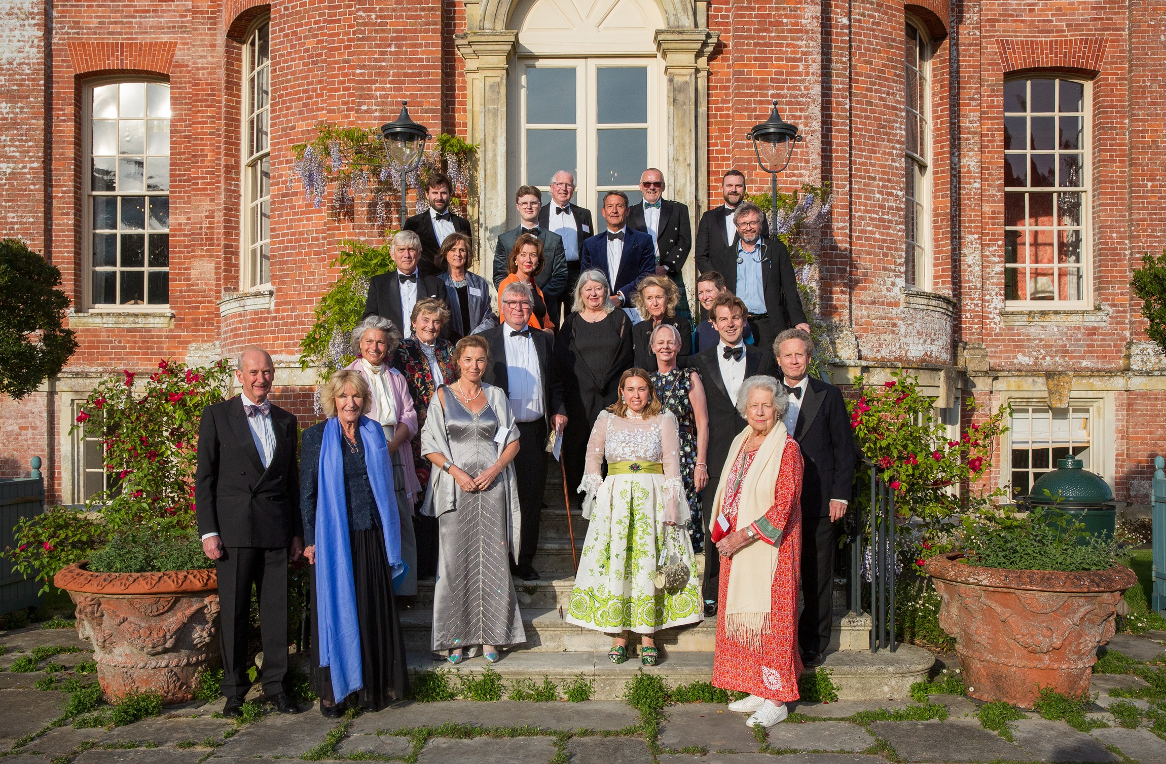 Formal group portrait of approximately 30 people in evening wear, including Annabel Elliot (Queen Camilla's sister) and other prominent Dorset residents, arranged in three rows in front of a red brick Georgian building with a white-columned central entrance. The group includes members of Dorset County Hospital Charity’s Emergency and Critical Care Appeal committee. Climbing wisteria and flowering plants frame the doorway, with terracotta urns positioned on either side of the steps. Photography by David Lewis.
