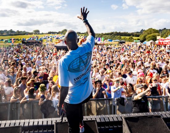 A performer on stage at an outdoor festival raises his arm to an enthusiastic crowd wearing a blue t-shirt. Thousands of festival-goers fill the field below, with multiple festival tents and marquees visible in the background, including a distinctive red and white striped circus-style tent. The rural setting features open countryside and blue sky with scattered clouds.
