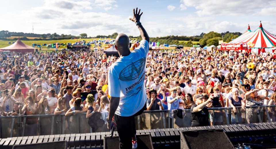 A performer on stage at an outdoor festival raises his arm to an enthusiastic crowd wearing a blue t-shirt. Thousands of festival-goers fill the field below, with multiple festival tents and marquees visible in the background, including a distinctive red and white striped circus-style tent. The rural setting features open countryside and blue sky with scattered clouds.