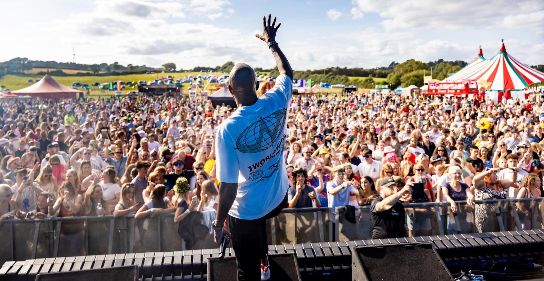 A performer on stage at an outdoor festival raises his arm to an enthusiastic crowd wearing a blue t-shirt. Thousands of festival-goers fill the field below, with multiple festival tents and marquees visible in the background, including a distinctive red and white striped circus-style tent. The rural setting features open countryside and blue sky with scattered clouds.