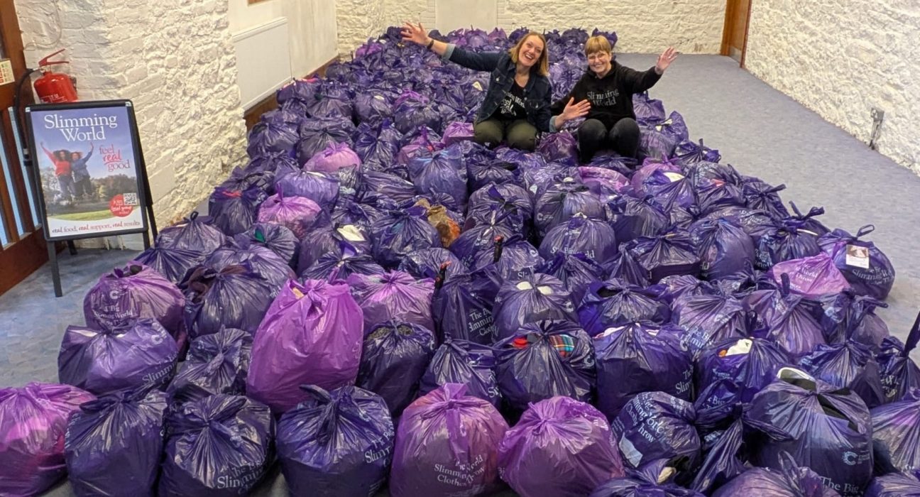 Two people sitting atop a large pile of purple and black plastic bags filled with donated clothing during a Slimming World clothes throw donation event. The bags are labeled "Slimming World Clothes Throw" and are stacked in a community space with white brick walls. A Slimming World promotional sign is visible on the left side of the room. A fire extinguisher is mounted on the wall.