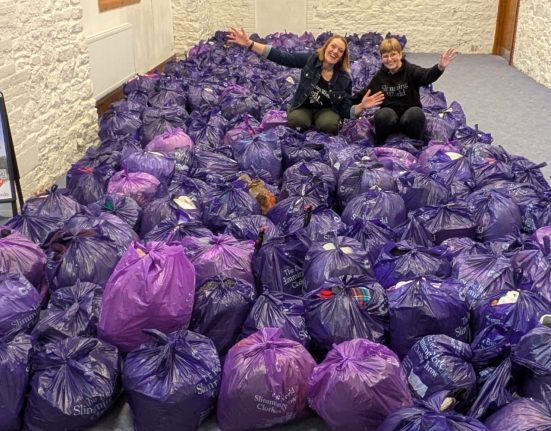 Two people sitting atop a large pile of purple and black plastic bags filled with donated clothing during a Slimming World clothes throw donation event. The bags are labeled "Slimming World Clothes Throw" and are stacked in a community space with white brick walls. A Slimming World promotional sign is visible on the left side of the room. A fire extinguisher is mounted on the wall.