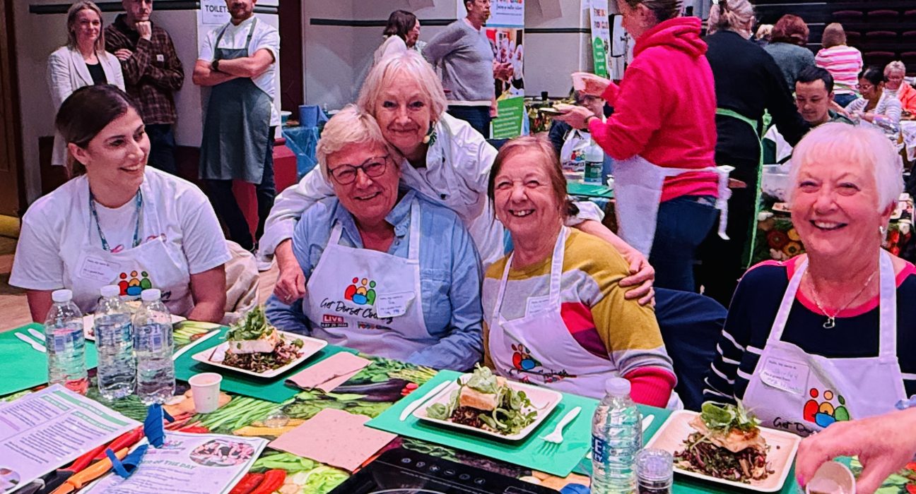 A group of people seated at a table during a community cooking event in a church hall with arched ceilings. The participants wear aprons with a colorful logo and are smiling while enjoying meals on green placemats. Plates of food, water glasses, and activity sheets are arranged on the table. Other participants and volunteers are visible in the background.