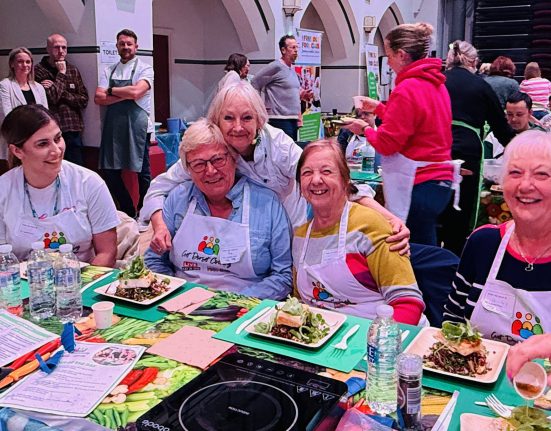 A group of people seated at a table during a community cooking event in a church hall with arched ceilings. The participants wear aprons with a colorful logo and are smiling while enjoying meals on green placemats. Plates of food, water glasses, and activity sheets are arranged on the table. Other participants and volunteers are visible in the background.