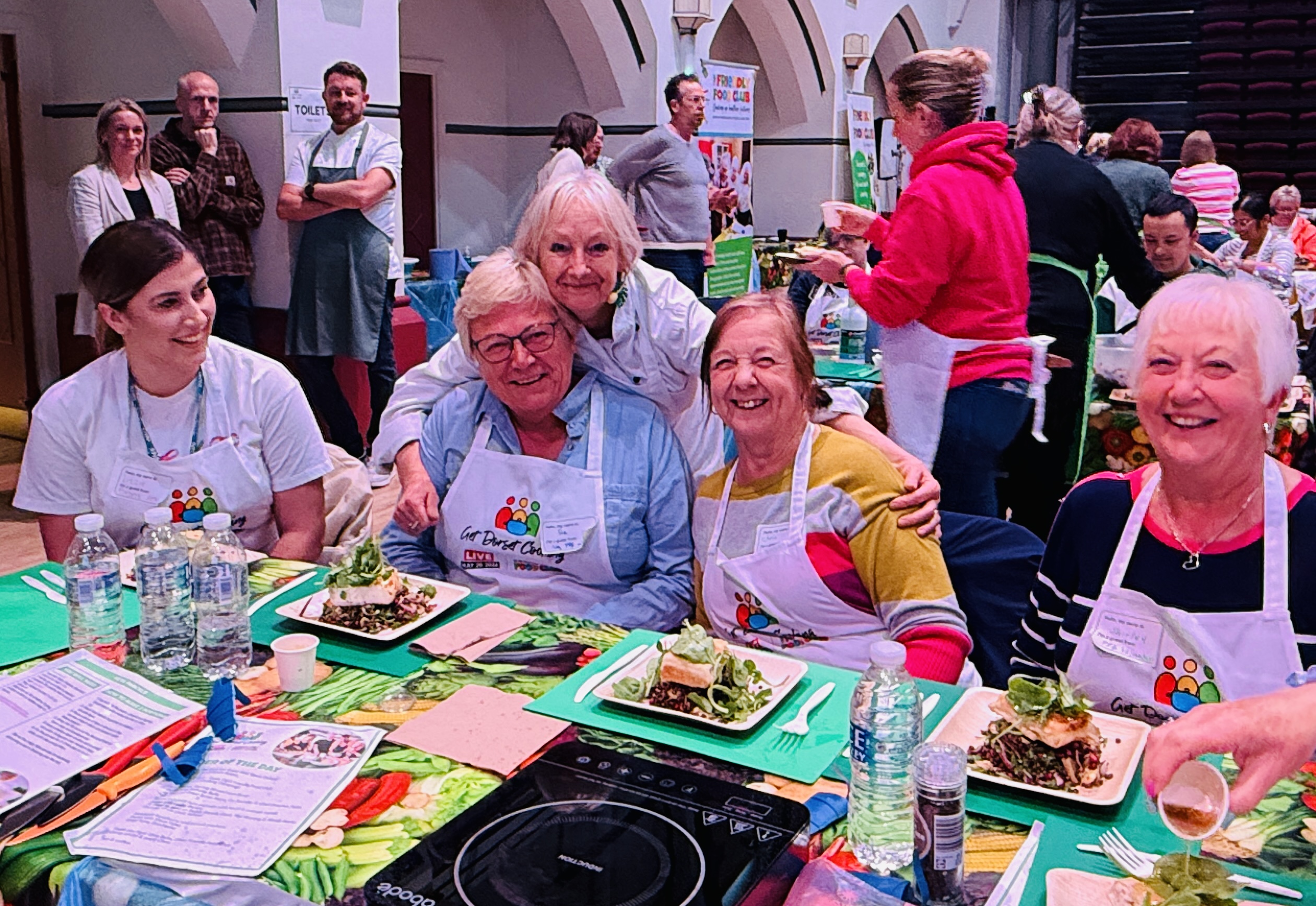 A group of people seated at a table during a community cooking event in a church hall with arched ceilings. The participants wear aprons with a colorful logo and are smiling while enjoying meals on green placemats. Plates of food, water glasses, and activity sheets are arranged on the table. Other participants and volunteers are visible in the background.