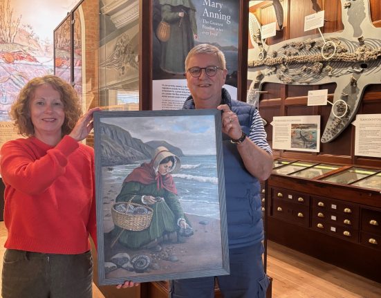 Bridget Houseago, Lyme Regis Museum director, and artist David Manners hold a framed painting depicting Mary Anning collecting fossils on Charmouth beach, set against coastal cliffs. They stand in the museum's exhibition space surrounded by geological specimens and fossil displays, with wooden display cabinets and museum artifacts visible in the background.