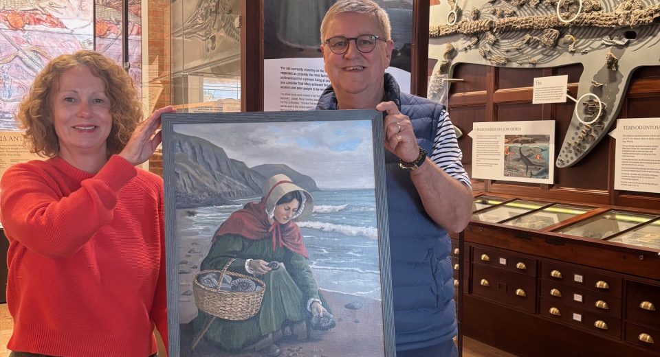 Bridget Houseago, Lyme Regis Museum director, and artist David Manners hold a framed painting depicting Mary Anning collecting fossils on Charmouth beach, set against coastal cliffs. They stand in the museum's exhibition space surrounded by geological specimens and fossil displays, with wooden display cabinets and museum artifacts visible in the background.