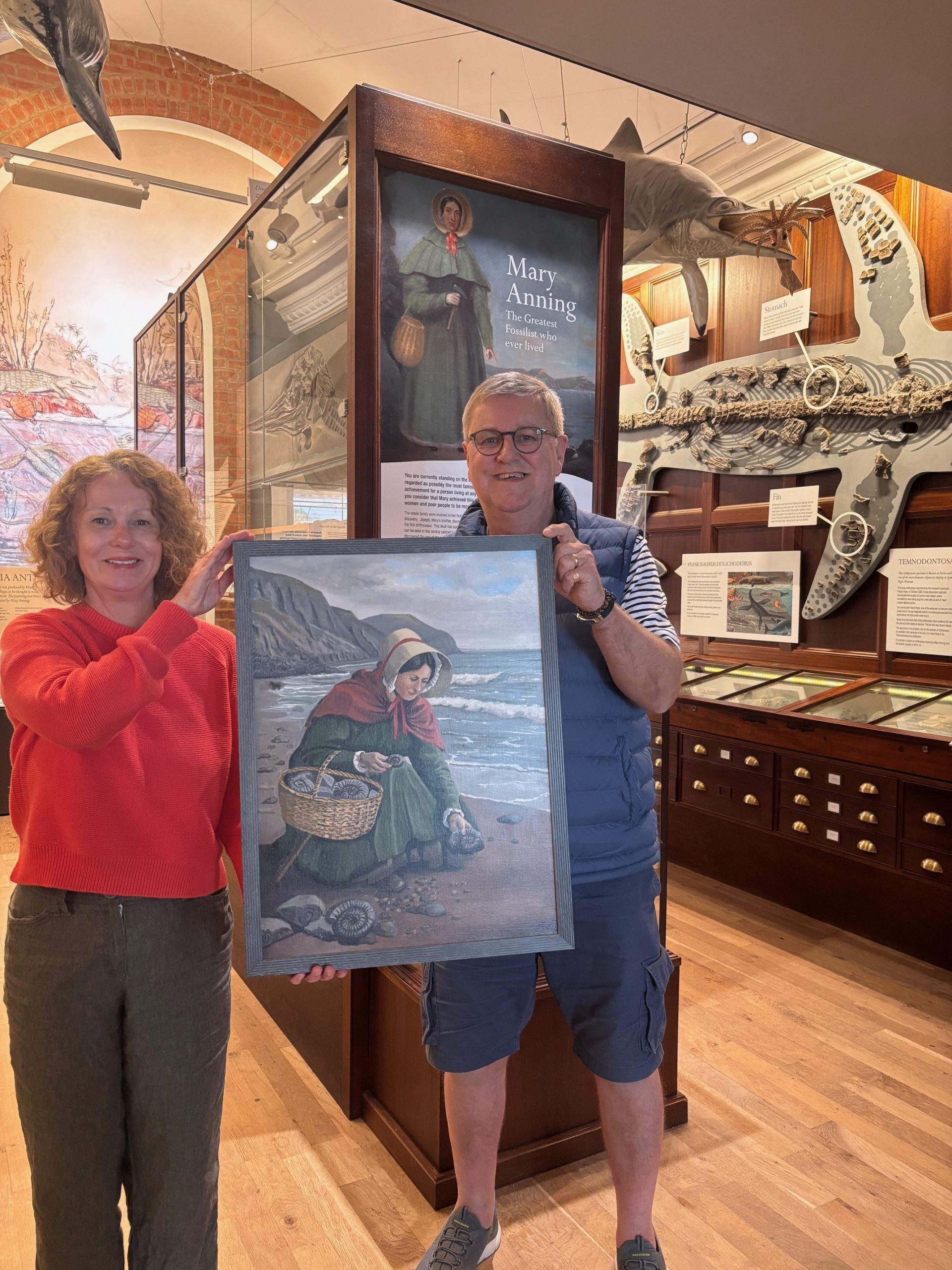 Bridget Houseago, Lyme Regis Museum director, and artist David Manners hold a framed painting depicting Mary Anning collecting fossils on Charmouth beach, set against coastal cliffs. They stand in the museum's exhibition space surrounded by geological specimens and fossil displays, with wooden display cabinets and museum artifacts visible in the background.