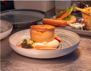 A gourmet plated dish featuring a golden-brown baked pastry case filled with creamy filling, served on a white ridged plate with roasted carrots, broccoli floret, and additional vegetables or garnish. The fine dining presentation includes similar plates visible in the background, on a dark wooden table.