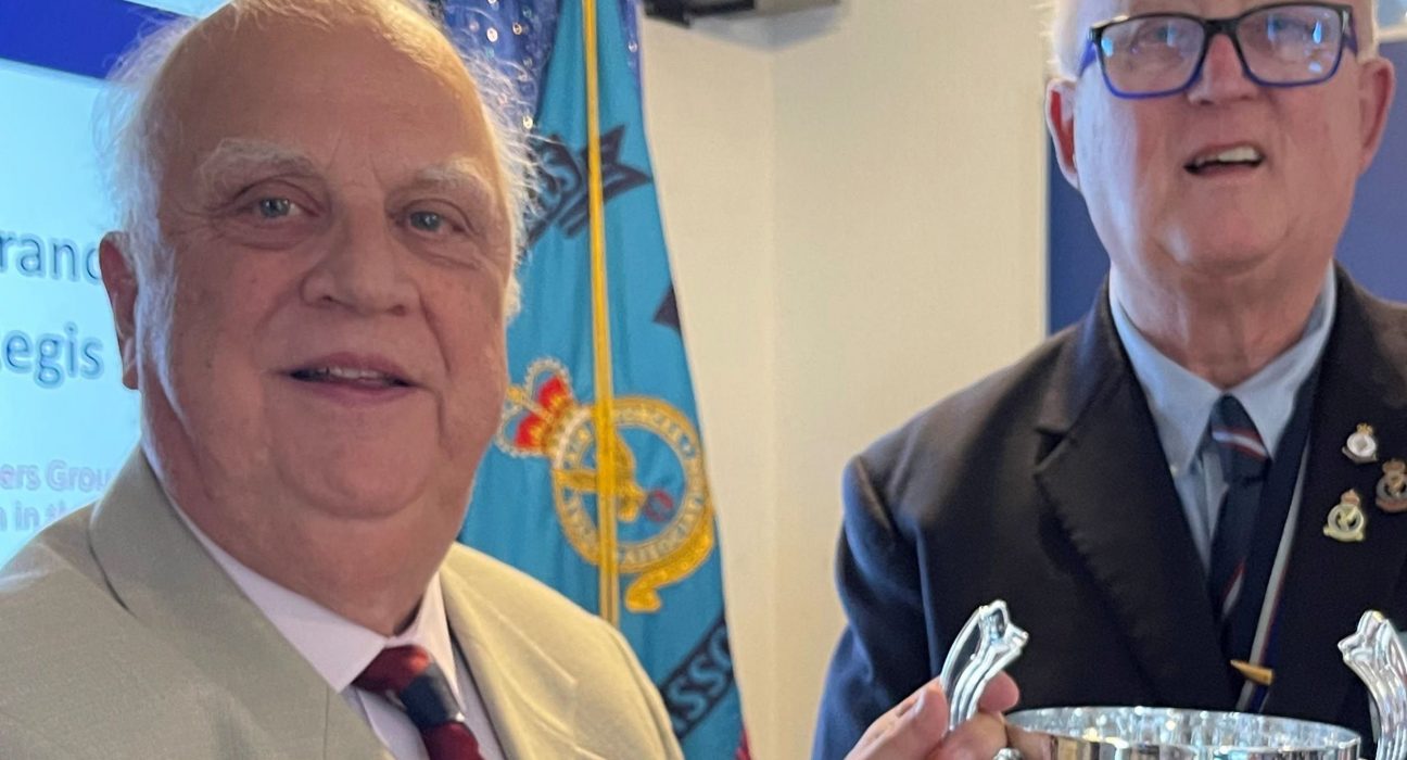 Two older men stand together at a Royal Air Forces Association (RAFA) event in front of a blue RAFA flag displaying the RAF emblem. The man on the left wears a light grey jacket with a burgundy and navy striped tie and RAF insignia pin. The man on the right wears a dark blue blazer with RAF medals and insignia on his chest and holds a polished silver trophy with decorative handles.