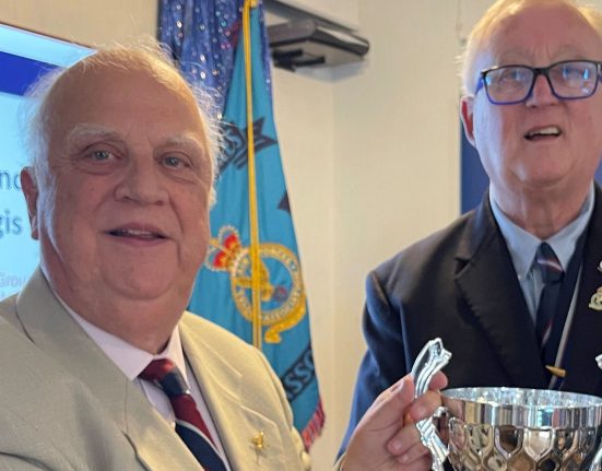 Two older men stand together at a Royal Air Forces Association (RAFA) event in front of a blue RAFA flag displaying the RAF emblem. The man on the left wears a light grey jacket with a burgundy and navy striped tie and RAF insignia pin. The man on the right wears a dark blue blazer with RAF medals and insignia on his chest and holds a polished silver trophy with decorative handles.