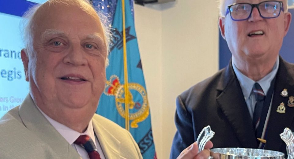 Two older men stand together at a Royal Air Forces Association (RAFA) event in front of a blue RAFA flag displaying the RAF emblem. The man on the left wears a light grey jacket with a burgundy and navy striped tie and RAF insignia pin. The man on the right wears a dark blue blazer with RAF medals and insignia on his chest and holds a polished silver trophy with decorative handles.