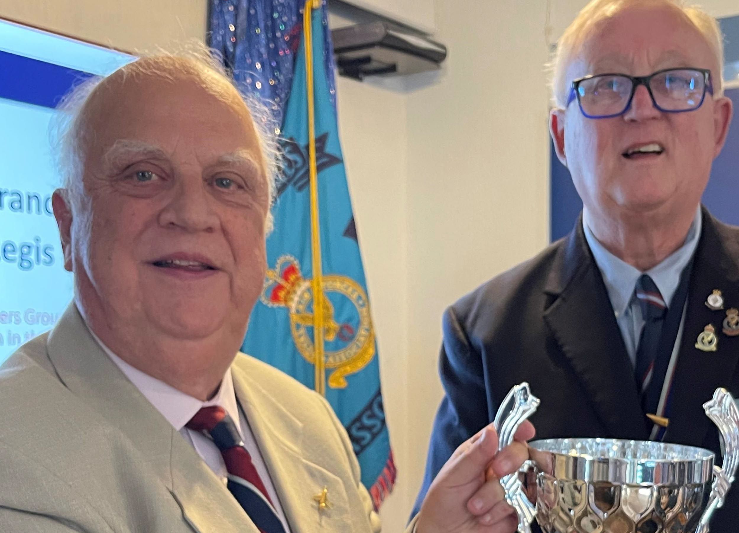 Two older men stand together at a Royal Air Forces Association (RAFA) event in front of a blue RAFA flag displaying the RAF emblem. The man on the left wears a light grey jacket with a burgundy and navy striped tie and RAF insignia pin. The man on the right wears a dark blue blazer with RAF medals and insignia on his chest and holds a polished silver trophy with decorative handles.