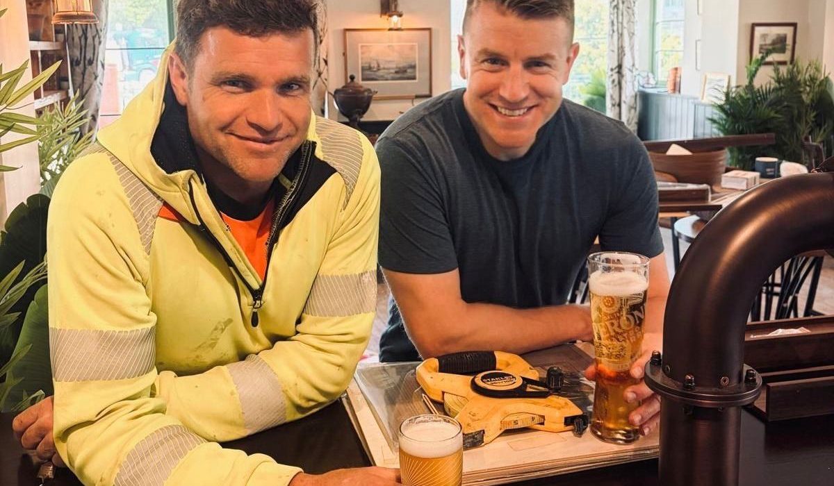 Two men sitting at a bar smiling at the camera. The man on the left wears a bright yellow jacket with an orange shirt and a lanyard badge, while the man on the right wears a dark navy t-shirt. Both hold pints of beer on the bar in front of them. Behind them is a warmly lit pub or lounge interior with green-trimmed walls, windows with patterned curtains, framed artwork, potted plants, and dark wood furniture.