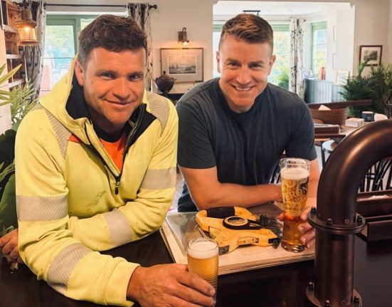 Two men sitting at a bar smiling at the camera. The man on the left wears a bright yellow jacket with an orange shirt and a lanyard badge, while the man on the right wears a dark navy t-shirt. Both hold pints of beer on the bar in front of them. Behind them is a warmly lit pub or lounge interior with green-trimmed walls, windows with patterned curtains, framed artwork, potted plants, and dark wood furniture.