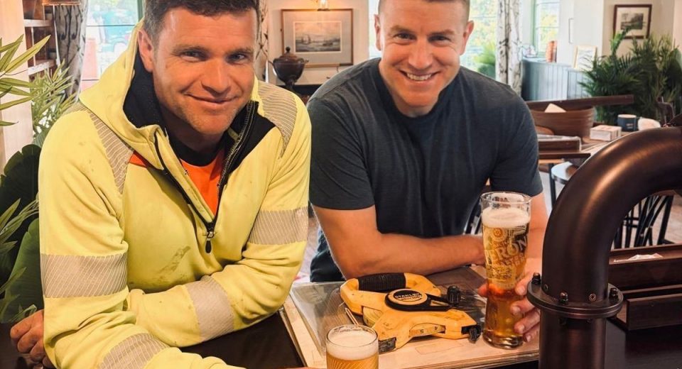 Two men sitting at a bar smiling at the camera. The man on the left wears a bright yellow jacket with an orange shirt and a lanyard badge, while the man on the right wears a dark navy t-shirt. Both hold pints of beer on the bar in front of them. Behind them is a warmly lit pub or lounge interior with green-trimmed walls, windows with patterned curtains, framed artwork, potted plants, and dark wood furniture.