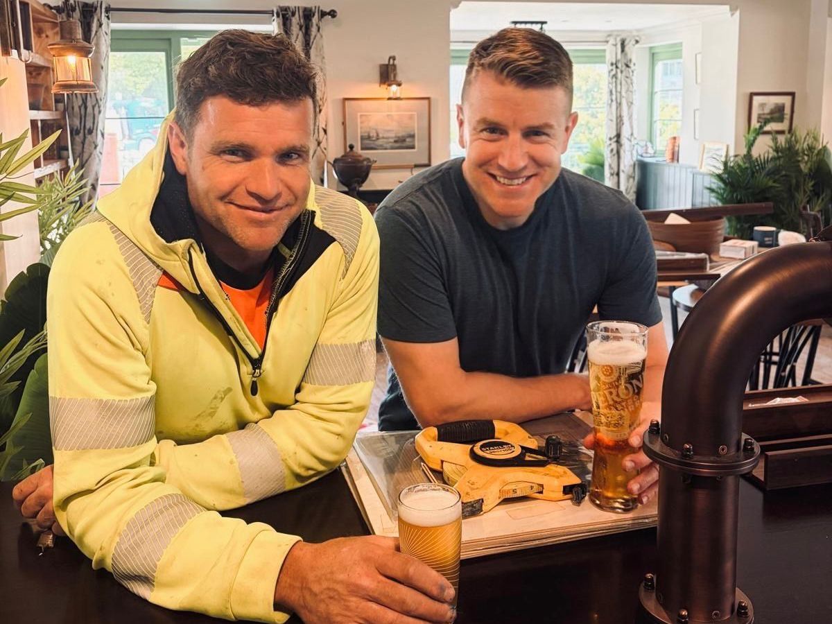 Two men sitting at a bar smiling at the camera. The man on the left wears a bright yellow jacket with an orange shirt and a lanyard badge, while the man on the right wears a dark navy t-shirt. Both hold pints of beer on the bar in front of them. Behind them is a warmly lit pub or lounge interior with green-trimmed walls, windows with patterned curtains, framed artwork, potted plants, and dark wood furniture.