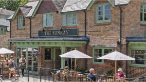 The Sunray pub with red brick facade and green-trimmed storefront, featuring outdoor patio seating with white umbrellas. Several patrons are seated at tables on the terrace, which is enclosed by black railings. The building has large windows and slate-colored roofing.