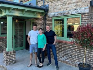 Three people smiling at the camera standing outside a brick storefront with mint-green painted trim and large windows. The man on the left wears a light blue t-shirt and shorts, the woman in the centre wears a turquoise top and black leggings, and the man on the right wears a dark navy t-shirt and jeans. A potted plant with burgundy foliage sits to the right of the entrance.