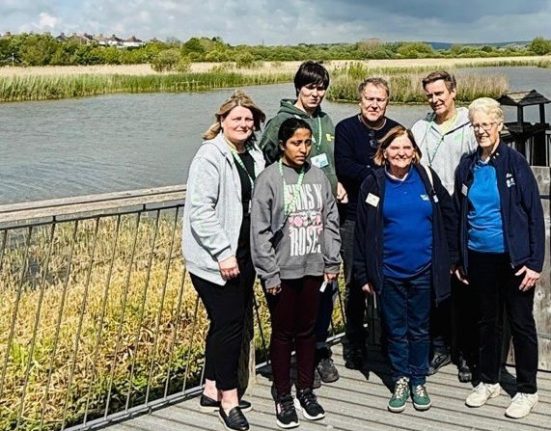 Seven people stand together on a wooden boardwalk or viewing platform overlooking a wetland nature reserve. The landscape behind them features tall reeds, open water, and tree lines under a partly cloudy sky. From left to right: a woman in a light cardigan and dark trousers, a young person in a dark hoodie, a man in a dark jacket with grey hair, a woman in a blue top with dark hair, a woman in a dark jacket in the centre, a man in light-coloured clothing, and an older woman in a dark jacket and blue top on the right.