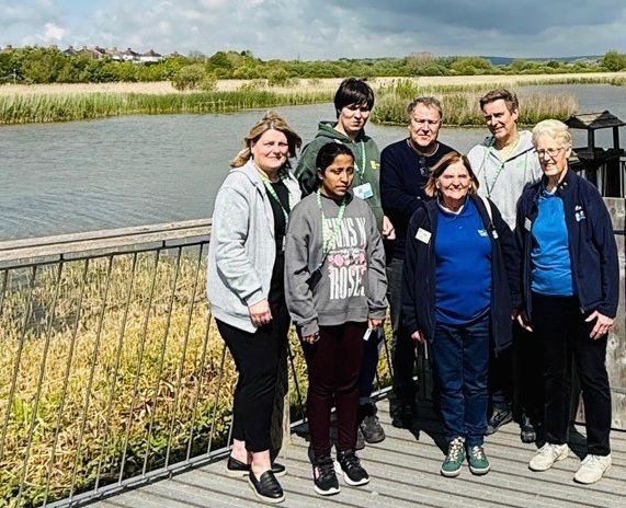 Seven people stand together on a wooden boardwalk or viewing platform overlooking a wetland nature reserve. The landscape behind them features tall reeds, open water, and tree lines under a partly cloudy sky. From left to right: a woman in a light cardigan and dark trousers, a young person in a dark hoodie, a man in a dark jacket with grey hair, a woman in a blue top with dark hair, a woman in a dark jacket in the centre, a man in light-coloured clothing, and an older woman in a dark jacket and blue top on the right.