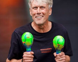 Bez from Happy Mondays is smiling at the camera wearing a black t-shirt with "MAHARISHI" text and holding two colorful maracas with green, blue, and orange spherical heads. The Jurassic Fields Festival logo is visible in the background.