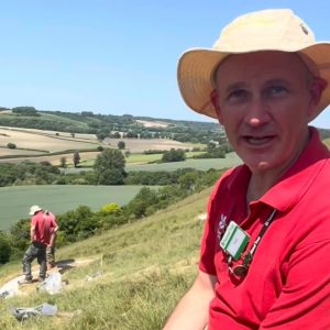 A National Trust volunteer in a red polo shirt, cream sun hat and name badge speaks to the camera on the hillside of the Cerne Abbas Giant, with the rolling Dorset countryside spread out behind him under a clear blue sky. Over his shoulder, another volunteer in matching red works on a freshly chalked white line of the Giant's outline, with tools and sacks of chalk lying on the grass nearby. The valley below is a patchwork of green and golden fields divided by hedgerows and clumps of trees, stretching away to wooded hills on the horizon in the summer sunshine.