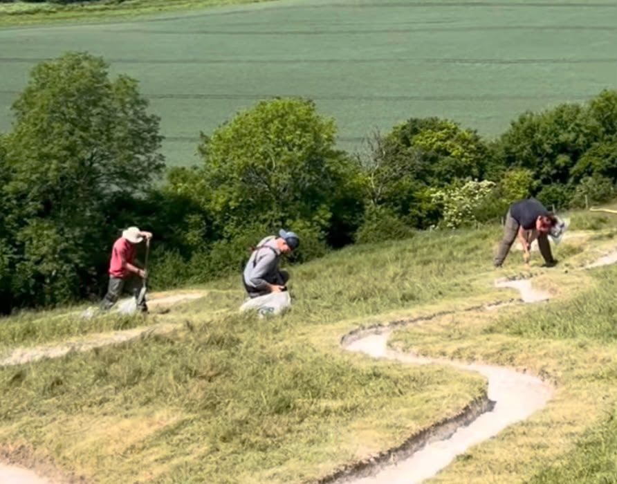 Three volunteers work on the steep grassy hillside of the Cerne Abbas Giant on a sunny day, re-chalking sections of the figure's outline. The chalk lines curve and wind across the slope in pale channels cut into the grass. On the left, a volunteer in a red top and wide-brimmed sun hat digs with a long-handled tool, while in the centre a man in a blue cap and grey top crouches beside a white sack of chalk. On the right, a woman in a dark top bends over the outline with a hand tool. Behind them, the Dorset countryside stretches out in a patchwork of green crop fields and mature trees under bright sunshine.