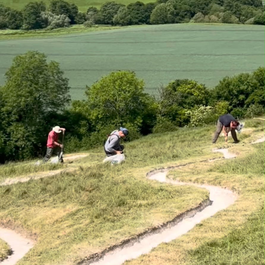 Three volunteers work on the steep grassy hillside of the Cerne Abbas Giant on a sunny day, re-chalking sections of the figure's outline. The chalk lines curve and wind across the slope in pale channels cut into the grass. On the left, a volunteer in a red top and wide-brimmed sun hat digs with a long-handled tool, while in the centre a man in a blue cap and grey top crouches beside a white sack of chalk. On the right, a woman in a dark top bends over the outline with a hand tool. Behind them, the Dorset countryside stretches out in a patchwork of green crop fields and mature trees under bright sunshine.