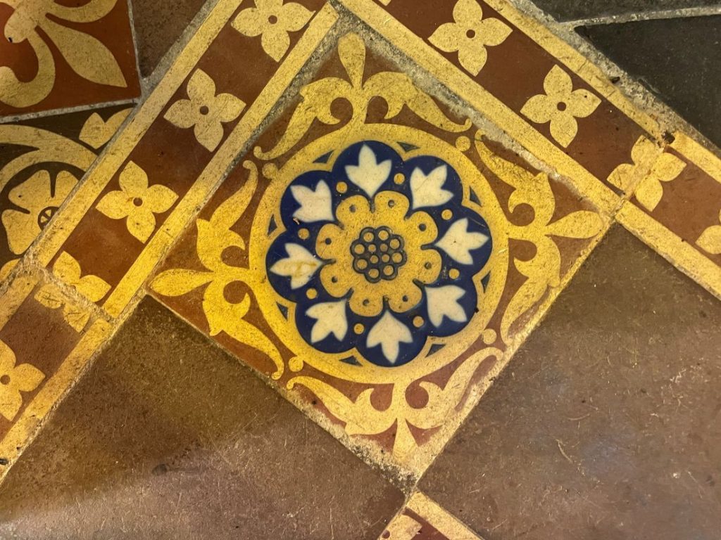 Close-up photograph of one of the Pugin-designed Minton encaustic tiles in the chancel of St Juthware Church. The square tile is set diagonally into the floor and features a central rosette design: a golden yellow flower with rounded petals sits inside a deep blue circle, with white heart-shaped leaves radiating outwards between the petals. Around the rosette, golden scrolling leaf patterns fill the corners of the tile against a terracotta red background. The tile is bordered by narrower strips decorated with small four-petalled yellow flowers, and plain brown tiles surround the design. The colours remain rich and vivid despite more than 150 years of footsteps, with light catching the glazed surface and showing the gentle wear of age.