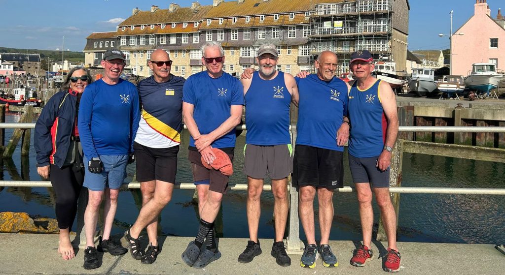 Seven members of Bridport Gig Rowing Club stand together on the quayside at West Bay Harbour, all wearing blue club shirts displaying the Bridport Gig Rowing Club logo. The group includes men and women of varying ages, mostly retired, wearing casual athletic wear and caps, with arms around each other's shoulders. Behind them are historic stone buildings including converted warehouses and a pink-painted building typical of the Dorset coast. The calm harbour waters and moored boats are visible, with the town's architecture rising behind. 
