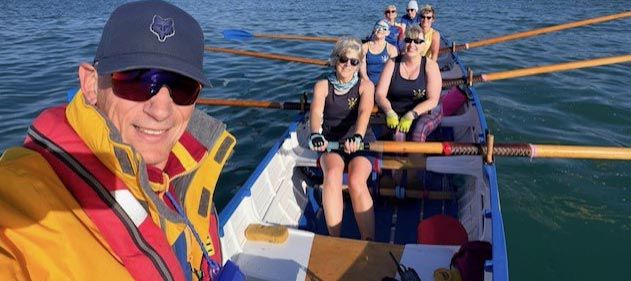 A group of Bridport Gig Rowing Club members row in a traditional wooden gig on calm water. In the foreground, a smiling rower wearing a yellow and red life jacket, dark cap and sunglasses holds an oar. Behind are four more rowers, predominantly women, wearing black sleeveless athletic tops and yellow life jackets, rowing in coordinated motion with wooden oars. The blue water and orange oars are visible, capturing a moment of camaraderie and enjoyment during their rowing session.