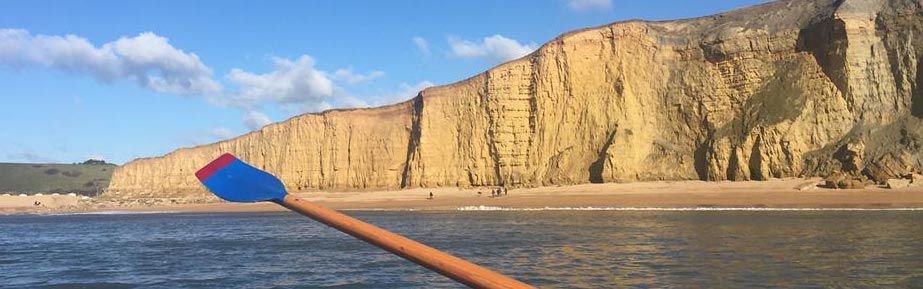 A close-up view of a wooden oar with a red and blue blade being held in the water, with West Bay's distinctive golden sandstone cliffs rising dramatically in the background beneath a clear blue sky. The cliffs show the characteristic layered rock formations that make West Bay a iconic Dorset landmark. The calm water of Lyme Bay stretches between the oar and the cliffs, capturing the serene beauty of the rowing location.