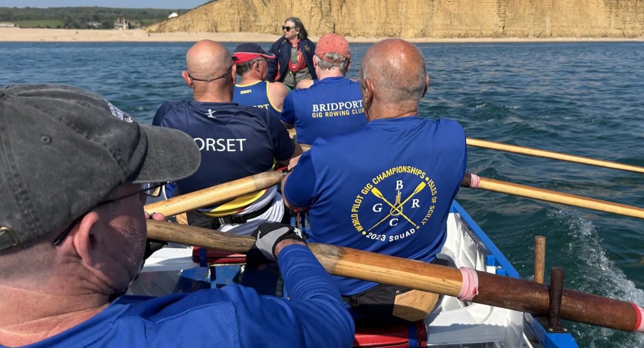 Members of Bridport Gig Rowing Club row in a traditional wooden Cornish pilot gig on Lyme Bay, pulling in practiced unison with wooden oars. The rowers wear blue club shirts and life jackets, with one shirt displaying "Bridport Gig Rowing Club" and another showing championship details. A cox in a red cap sits in the centre directing the crew. The golden cliffs of West Bay rise dramatically in the background beneath a clear blue sky. The image captures the dynamic movement and teamwork of competitive gig rowing.