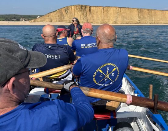Members of Bridport Gig Rowing Club row in a traditional wooden Cornish pilot gig on Lyme Bay, pulling in practiced unison with wooden oars. The rowers wear blue club shirts and life jackets, with one shirt displaying "Bridport Gig Rowing Club" and another showing championship details. A cox in a red cap sits in the centre directing the crew. The golden cliffs of West Bay rise dramatically in the background beneath a clear blue sky. The image captures the dynamic movement and teamwork of competitive gig rowing.