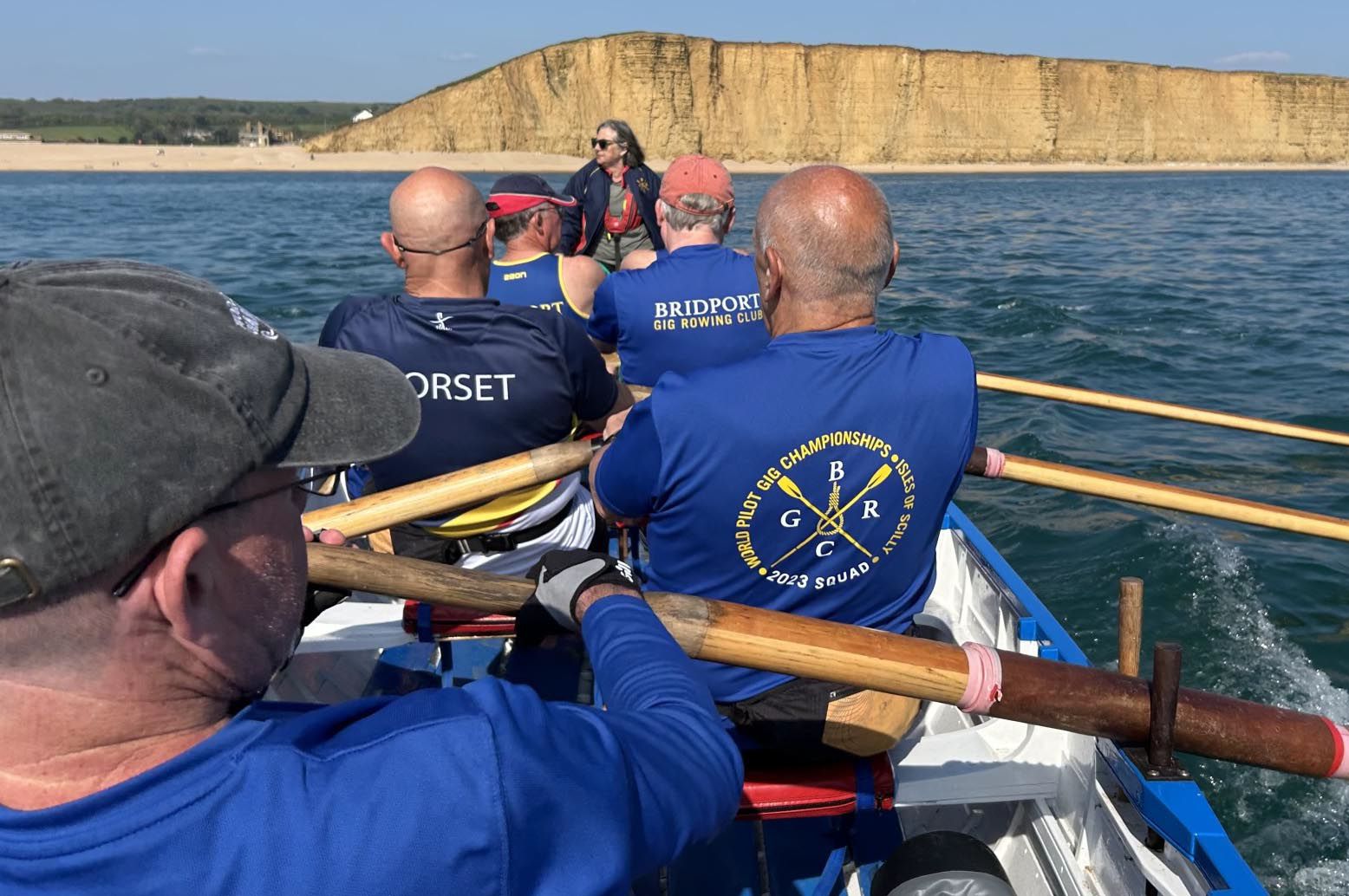 Members of Bridport Gig Rowing Club row in a traditional wooden Cornish pilot gig on Lyme Bay, pulling in practiced unison with wooden oars. The rowers wear blue club shirts and life jackets, with one shirt displaying "Bridport Gig Rowing Club" and another showing championship details. A cox in a red cap sits in the centre directing the crew. The golden cliffs of West Bay rise dramatically in the background beneath a clear blue sky. The image captures the dynamic movement and teamwork of competitive gig rowing.