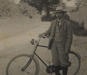 Sepia-toned historical photograph of Thomas Hardy, the renowned Dorset novelist and poet, standing beside a vintage bicycle on a tree-lined country road. He wears a formal suit jacket, patterned trousers, waistcoat and fedora hat. The photograph dates from the early-to-mid 20th century. Trees and foliage are visible in the soft-focused background.
