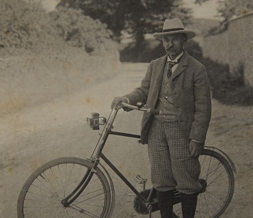 Sepia-toned historical photograph of Thomas Hardy, the renowned Dorset novelist and poet, standing beside a vintage bicycle on a tree-lined country road. He wears a formal suit jacket, patterned trousers, waistcoat and fedora hat. The photograph dates from the early-to-mid 20th century. Trees and foliage are visible in the soft-focused background.